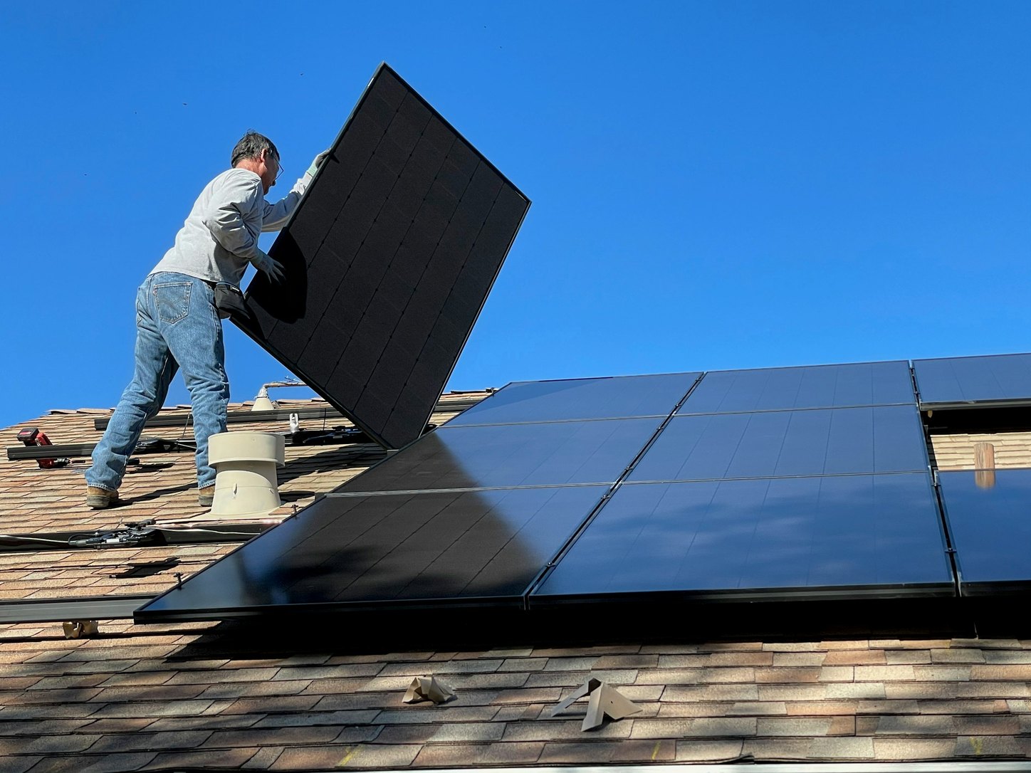 An installer lifting a black photovoltaic module into place on a shingled residential roof under a clear sky, beside a completed row of panels